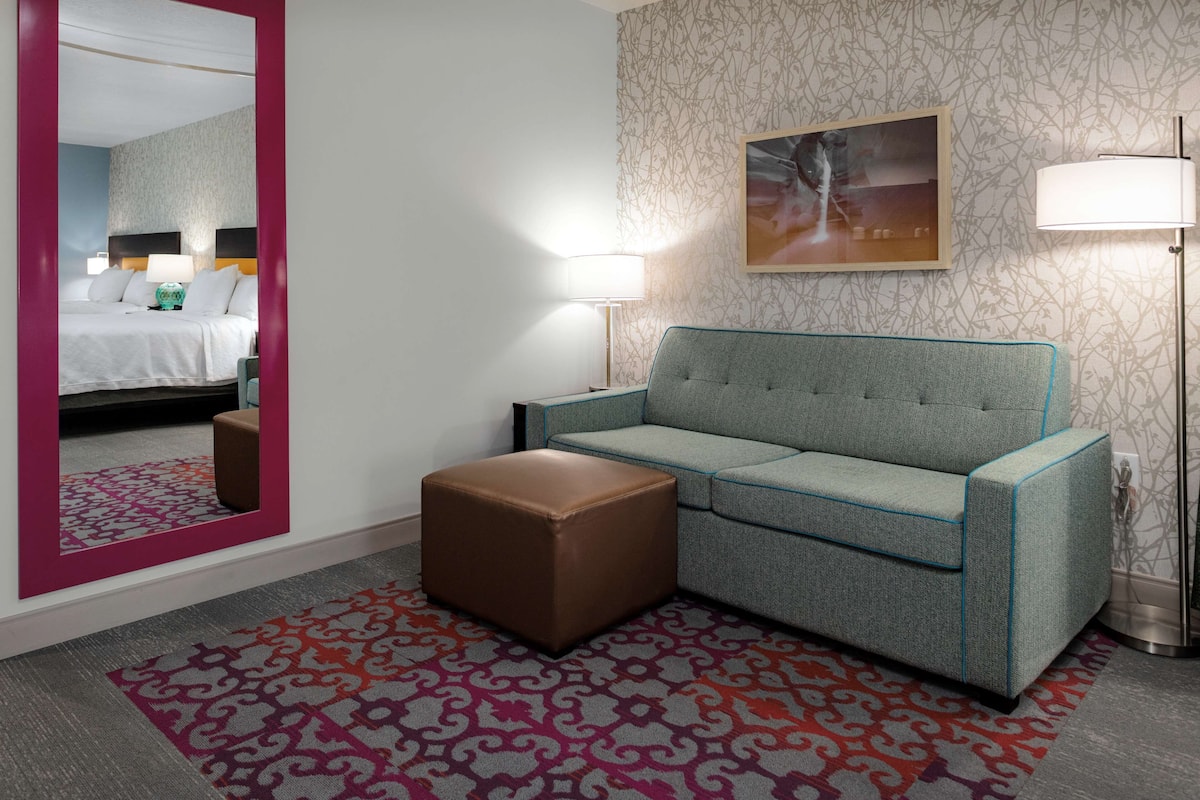 Una habitación de hotel con una cama, un sofá, un pequeño banquillo, una alfombra estampada y una pared con un cuadro enmarcado.