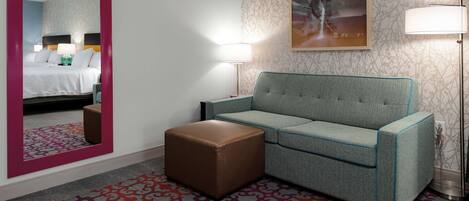 A hotel room with a bed, a sofa, a small ottoman, a patterned rug, and a wall with a framed picture.