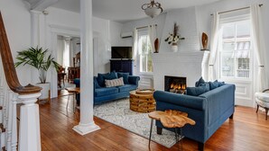 A spacious living room with a fireplace, blue sofas, wooden flooring, and a large window with curtains. - The Ashley Rose (Cape May)