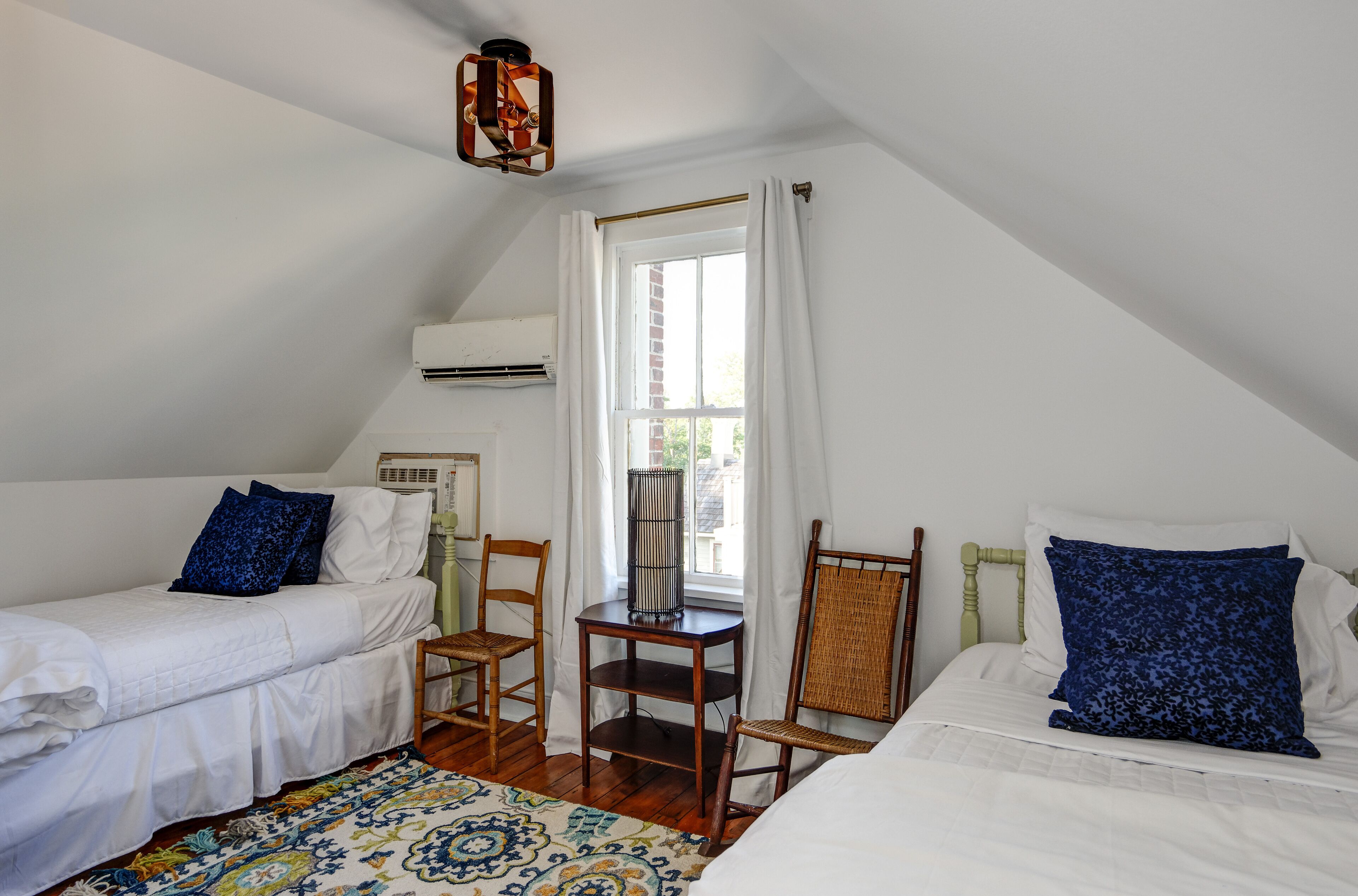 Een slaapkamer met twee eenpersoonsbedden, een klein tafeltje, twee stoelen, een raam met gordijnen en een airconditioning.
