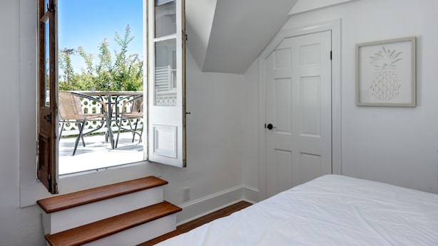 A bedroom with a large window, a wooden bench, a bed, and a door. - The Beach Rose (Cape May)