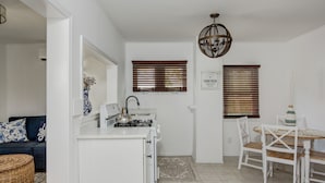 A compact kitchen with a white countertop, a stove, and a sink. There is a dining table with chairs and a decorative vase. A wicker basket is on the floor, and a blue and white patterned pillow is on a dark sofa. - The Beach Rose (Cape May)