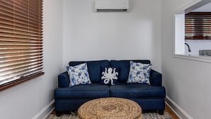 A blue sofa with patterned cushions, a woven ottoman, and a window with blinds. - The Beach Rose (Cape May)