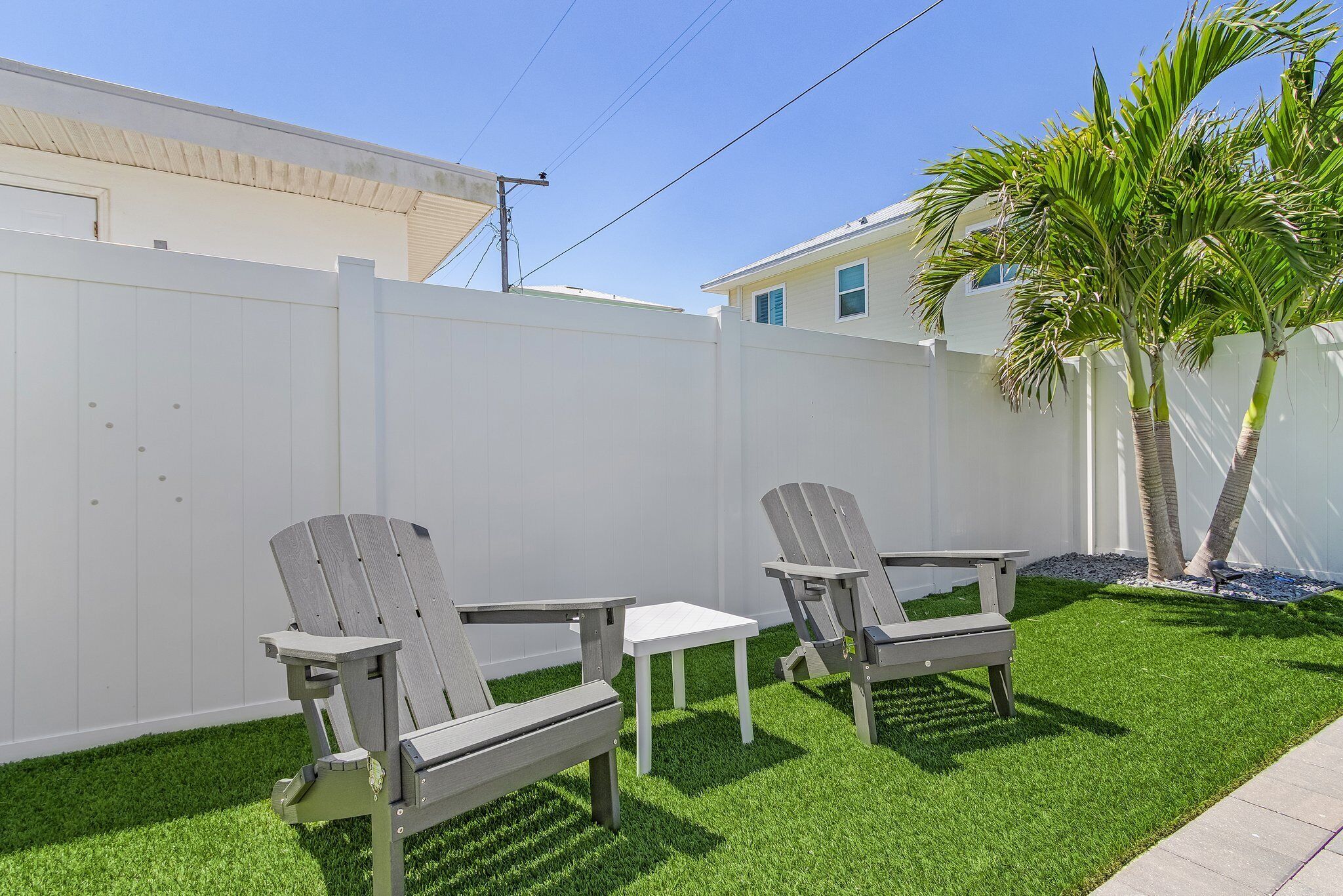 Un jardín trasero con césped artificial, dos sillas Adirondack, una mesita y una valla blanca.