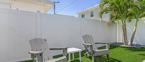 Un jardín trasero con césped artificial, dos sillas Adirondack, una mesita y una valla blanca.