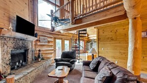A log cabin interior with a stone fireplace, leather furniture, a wooden staircase, and a large window. - Star Views - 2BR/New Hot Tub/New Firepit/Grill/Sleeps 8 (Gatlinburg)