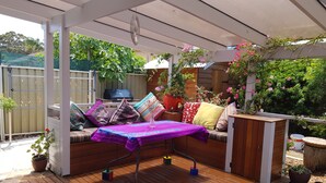 Ein überdachter Außenbereich mit Holzterrasse, gepolsterten Sitzmöglichkeiten und einem Tisch mit lila Tischdecke.