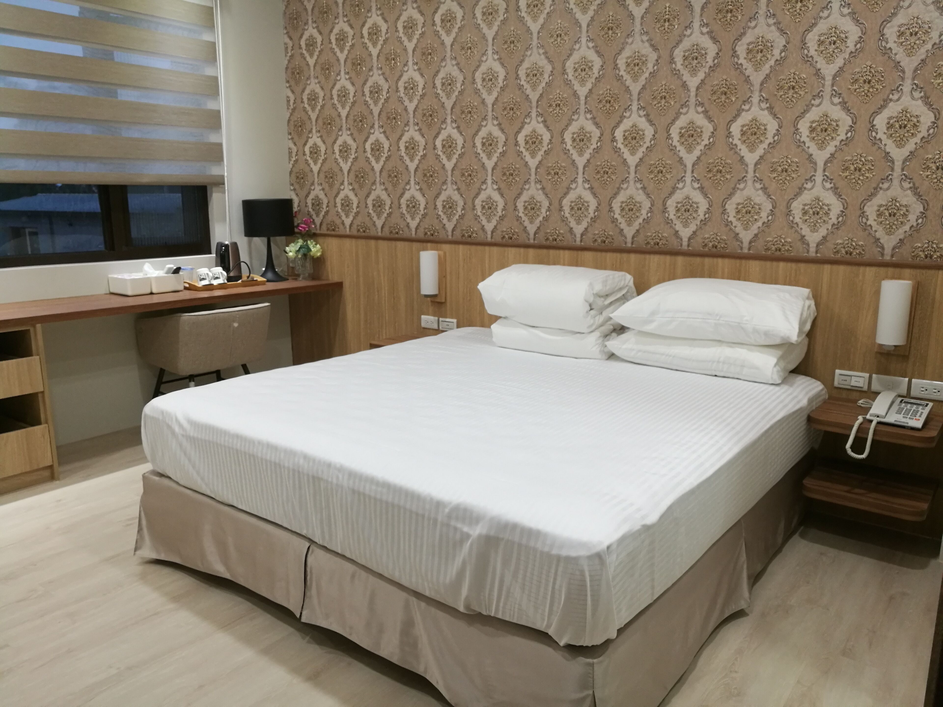 Habitación de hotel con una cama grande, un escritorio, una silla, una mesita de noche con teléfono y una pared con papel tapiz estampado.
