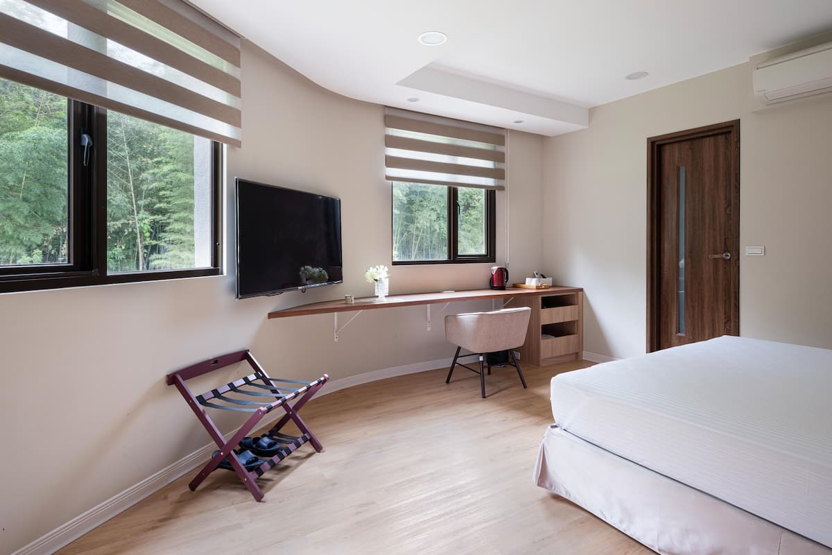A bedroom with a bed, a desk with a chair, a TV, and a window with blinds.