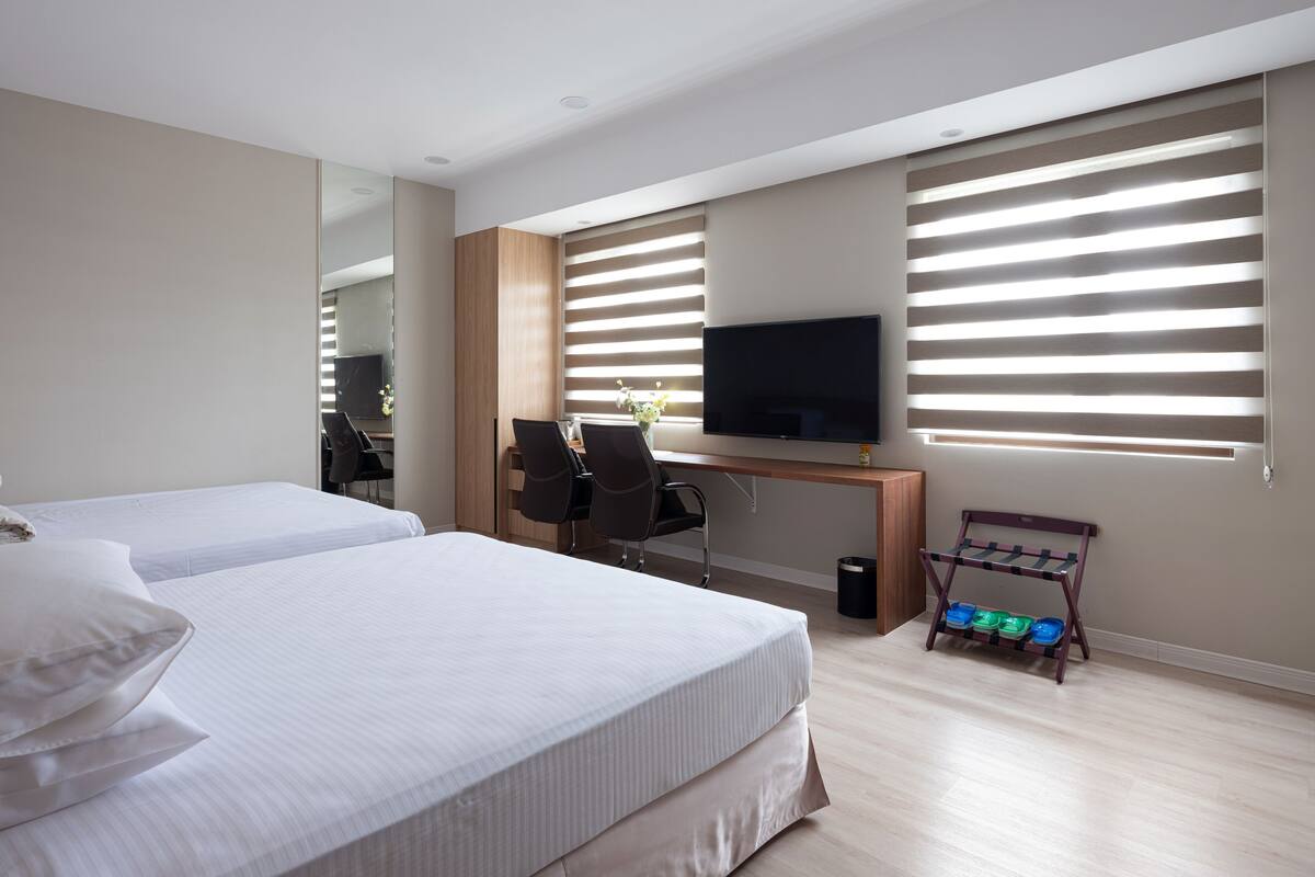 A hotel room with two beds, a desk, a TV, and a window with blinds.