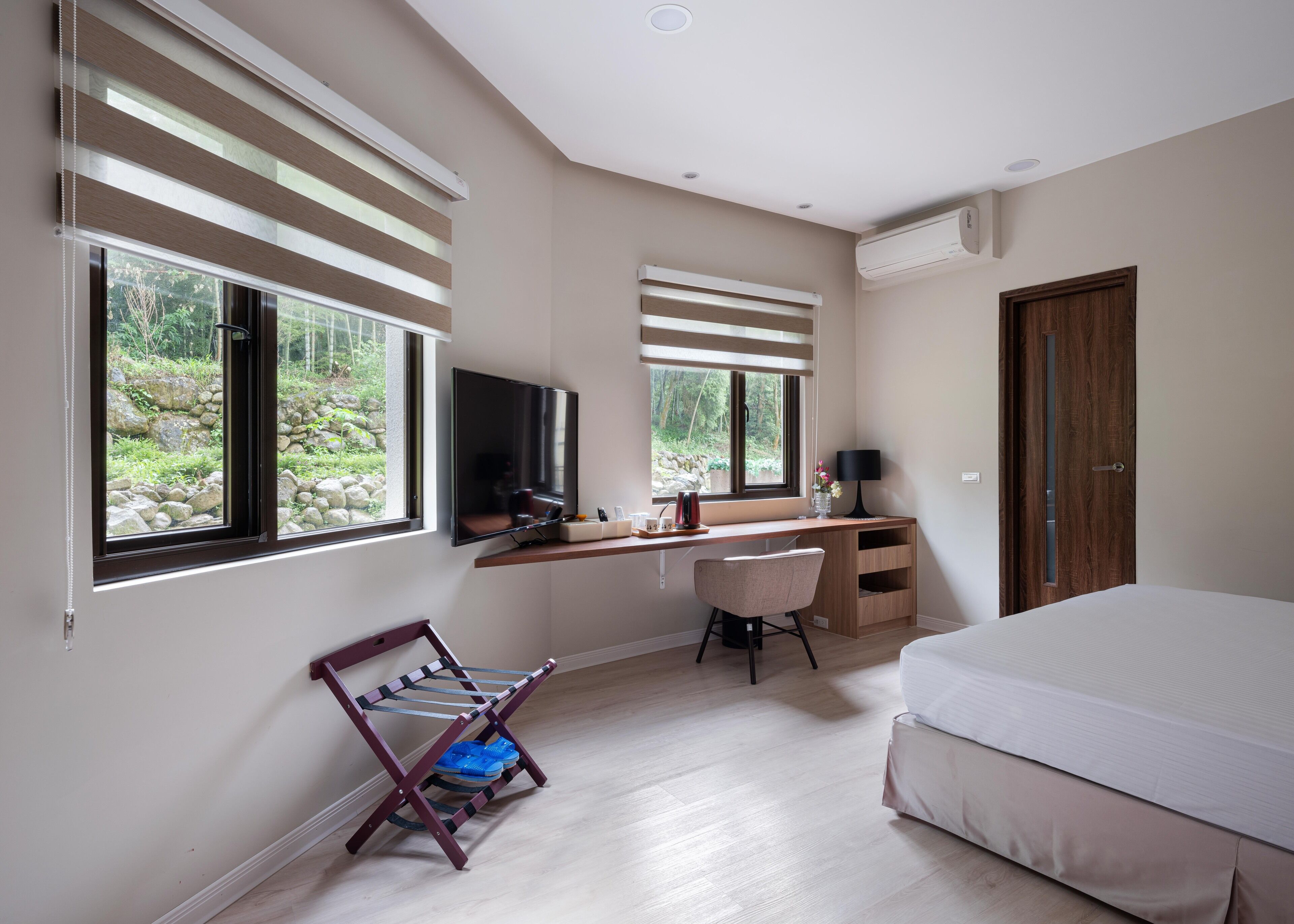 A bedroom with a bed, a desk with a chair, a TV, and a window with blinds.