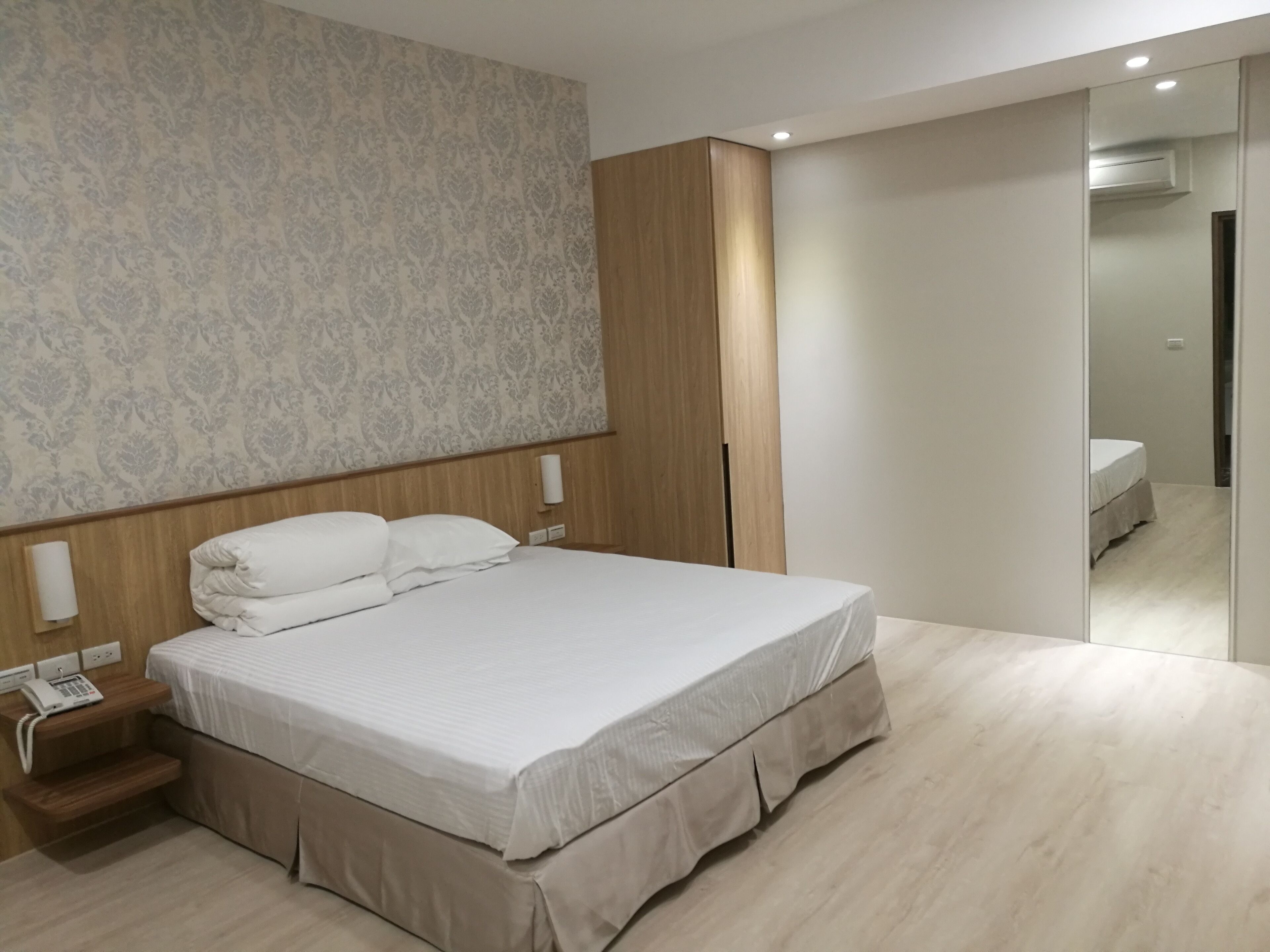 Una habitación de hotel moderna con una cama grande, un armario de madera, un teléfono en la mesita de noche y un espejo.