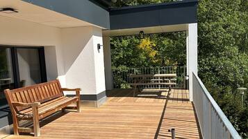Une terrasse en bois, agrémentée d’un banc et d’une table, entourée d’une balustrade et donnant sur un jardin planté d’arbres.