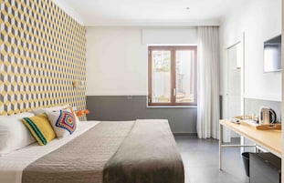 Quarto de hotel com cama, escrivaninha, janela com cortinas e parede estampada.