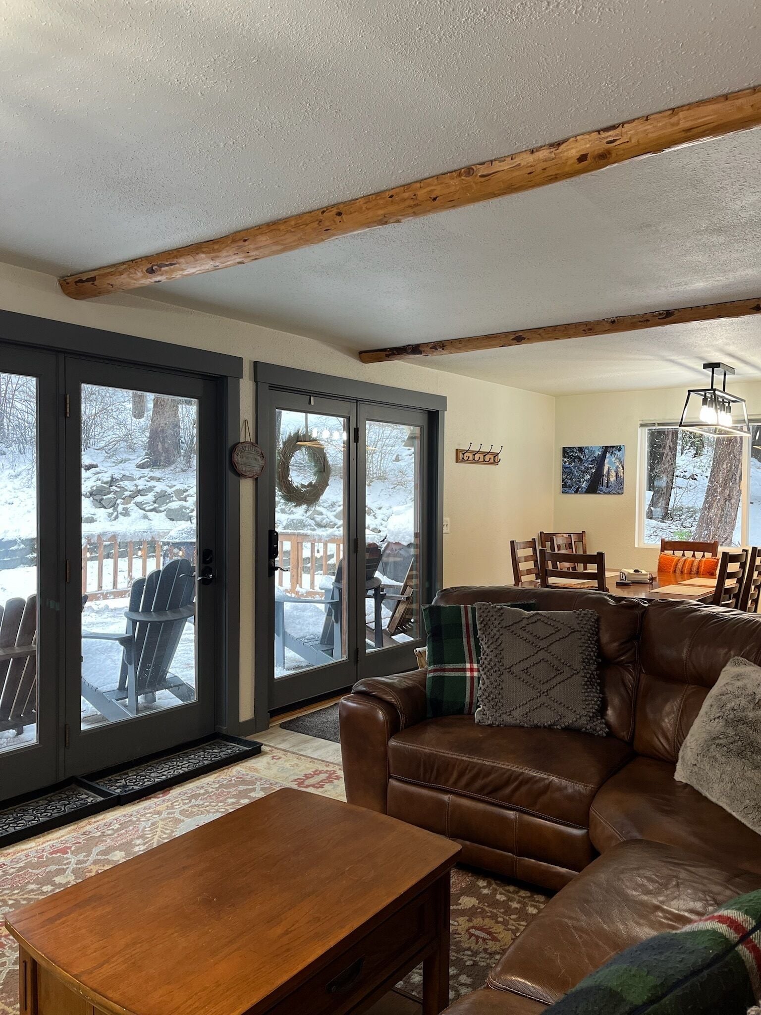 A cozy living room with a brown leather couch, a wooden coffee table, and a large window overlooking a snowy deck with Adirondack chairs.