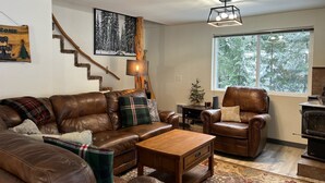 A cozy living room with a brown leather sofa, a wooden coffee table, a plaid throw pillow, a fireplace, and a large window with a view of trees.