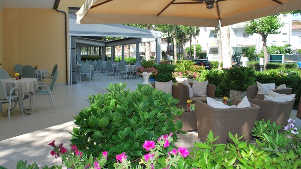 An outdoor patio area with tables and chairs, surrounded by greenery and flowers. - Hotel Cristoforo Colombo (Cesenatico)