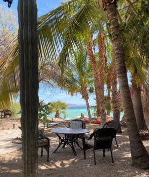 Un espacio de descanso frente a la playa, con una mesa y sillas, rodeado de palmeras y vistas al océano.