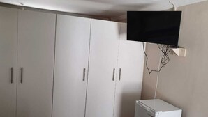 A room with white wardrobes, a flat-screen TV mounted on the wall, and a white refrigerator. - The Monument Guesthouse (Gqeberha)