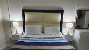 A hotel room with a large bed, two bedside tables, a nightstand, a mirror, and a small refrigerator. - The Monument Guesthouse (Gqeberha)