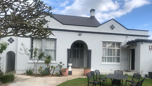 A white house with a grey roof, a front door, and a small patio with a table and chairs. - The Monument Guesthouse (Gqeberha)