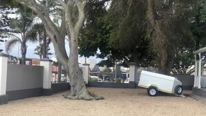 A parking area with a white trailer, a large tree, and a building with a grey facade. - The Monument Guesthouse (Gqeberha)