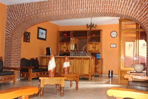 A cozy living room with a brick archway, wooden furniture, a dining table, and a kitchen area with cabinets and a clock. - Hotel Camino de Villaseca (Guanajuato)