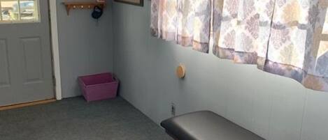 A room with a bench, a window with patterned curtains, a door, a small shelf with items, and a framed picture on the wall.