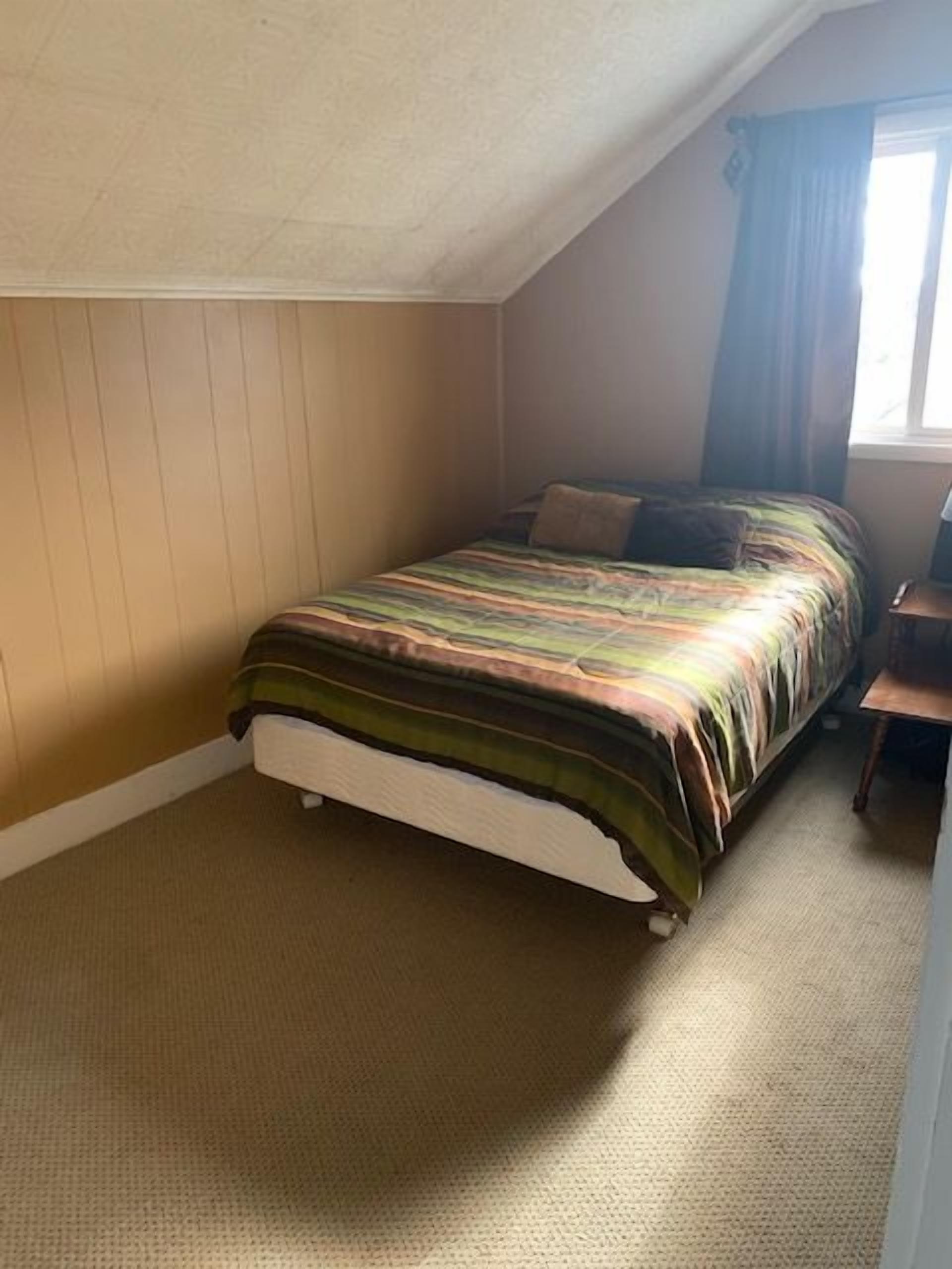 A bedroom with a single bed, a wooden headboard, a small bedside table, and a window with curtains.