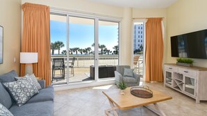 Un salon moderne avec un canapé, une table basse et un téléviseur. Il est doté de grandes portes-fenêtres coulissantes donnant sur un balcon avec vue sur des palmiers et une plage.