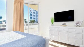Une chambre avec un grand lit, un téléviseur posé sur une commode blanche et un balcon offrant une vue sur un immeuble et des palmiers.