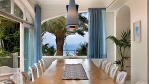 Ein langer Holztisch mit Stühlen, Blick auf das Meer und ein großes Fenster mit Vorhängen.