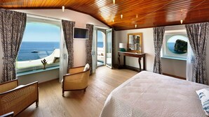 Ein geräumiges Zimmer mit einem großen Bett, einem Sessel, einem kleinen Tisch und einem Fenster mit Meerblick.