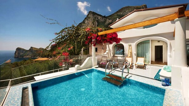 Ein Pool mit Blick auf das Meer und die Berge, umgeben von einem weißen Gebäude mit Balkon und Liegestühlen.
