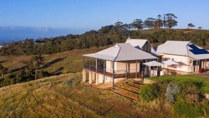 A modern house with a large glass enclosure, surrounded by a hilly landscape and trees.