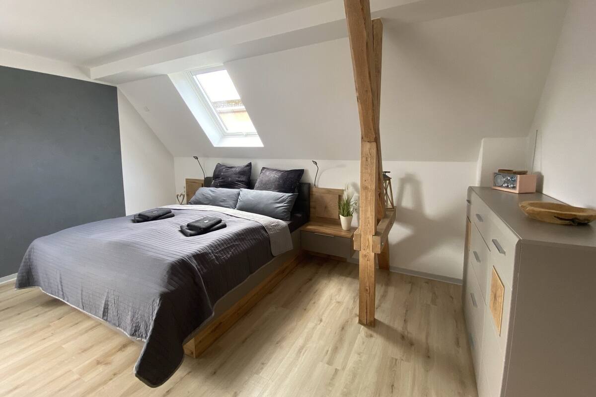 A modern bedroom with a sloped ceiling, a skylight, a bed with a grey and black bedspread, a wooden nightstand, and a small table with a radio.