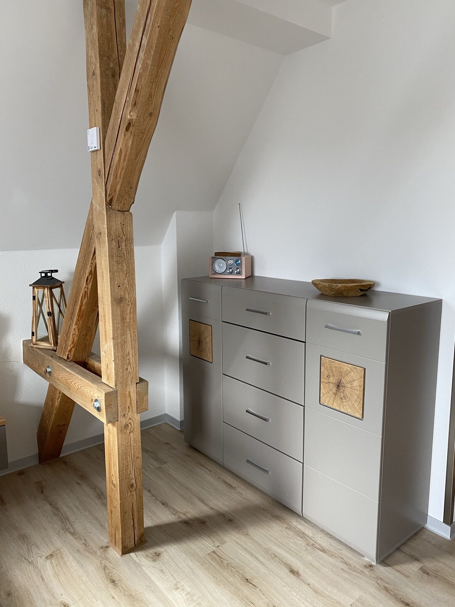 Una habitación con estructura de vigas de madera, un mueble gris con cajones, una radio y una pequeña mesa con un cuenco.