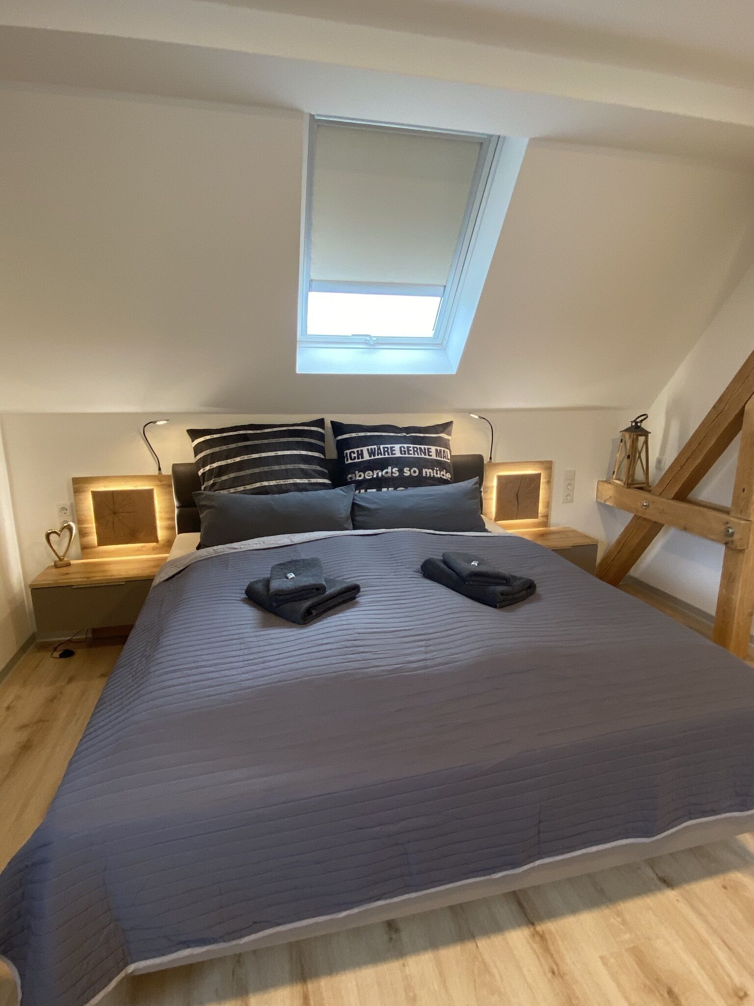 Une chambre avec un plafond en pente, une lucarne, un lit recouvert d’une couverture bleu foncé, deux tables de chevet avec des lampes et une structure en forme d’échelle en bois.