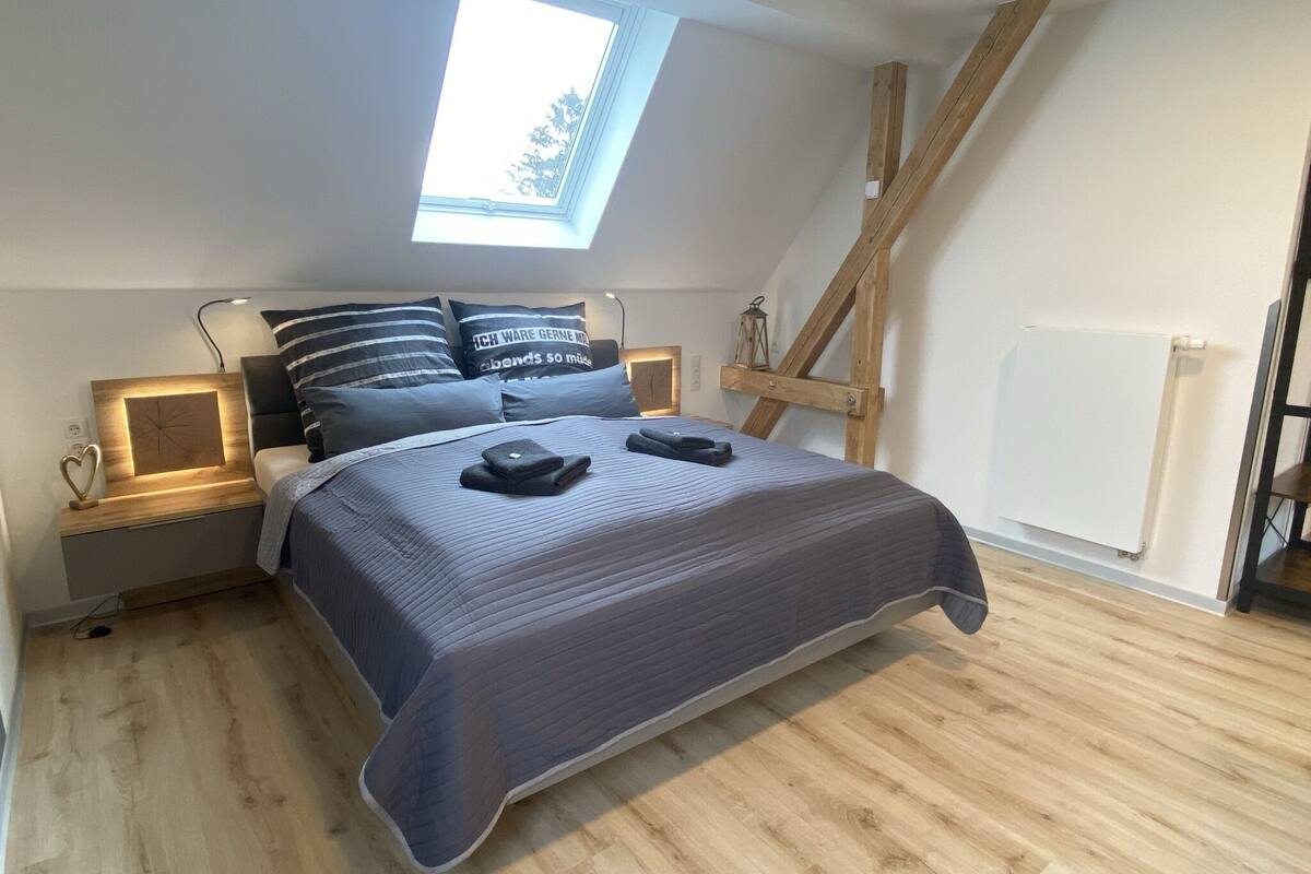 A bedroom with a skylight, wooden beams, a bed with a dark blue bedspread, and a nightstand with a lamp.