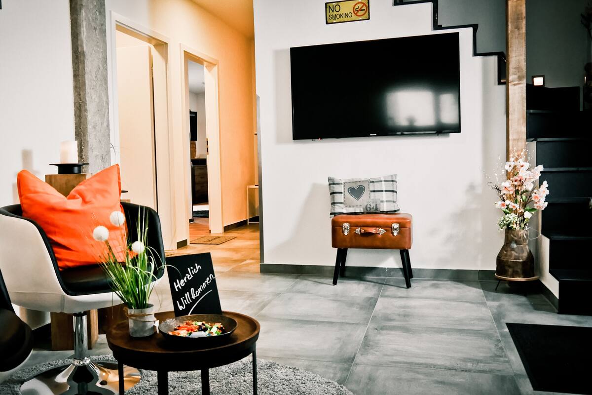 A modern living room with a sofa, a coffee table with a sign, a TV mounted on the wall, and a staircase.