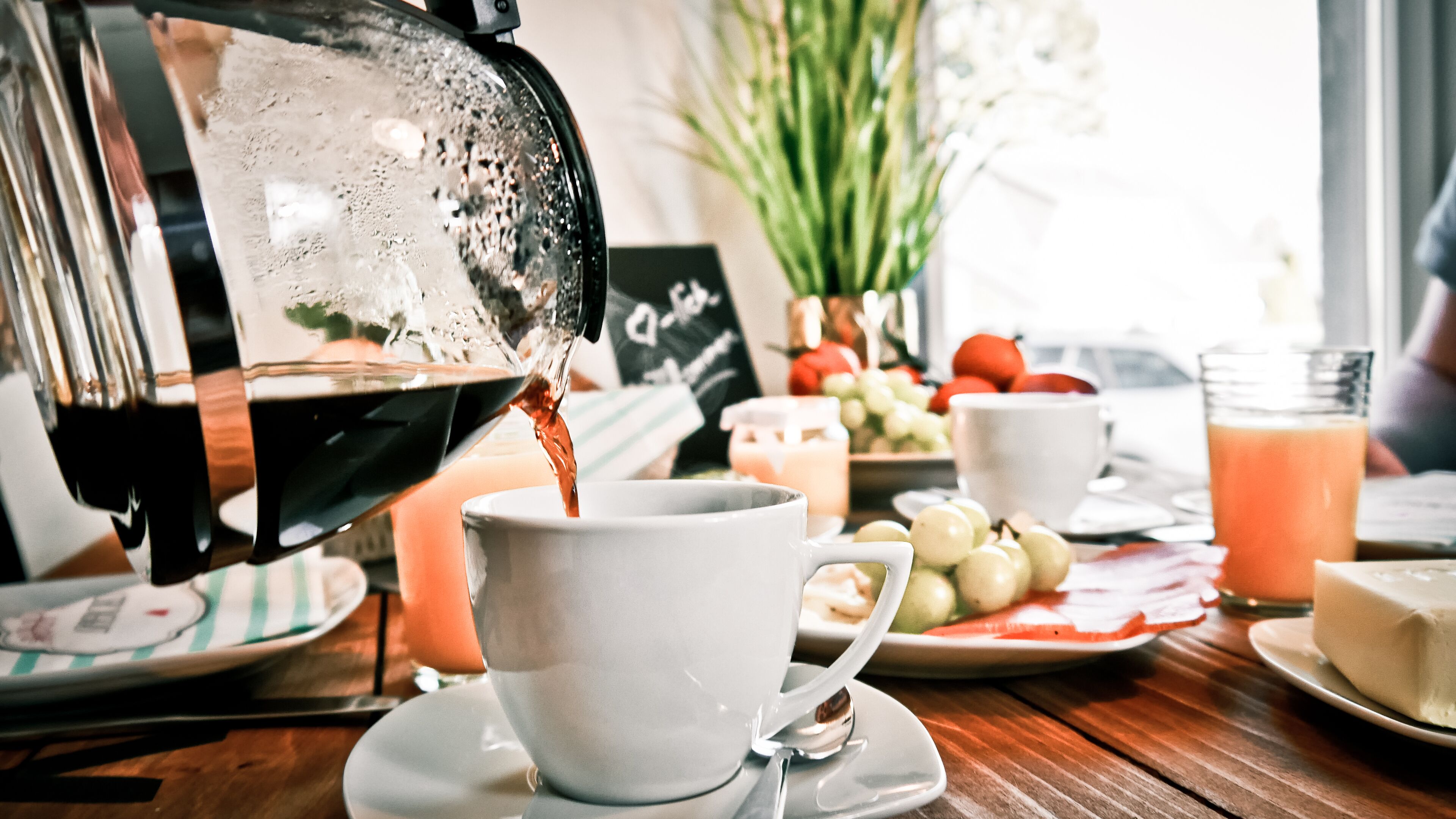 A coffee machine pouring coffee into a cup, a plate with grapes, a glass of orange juice, and a vase with flowers on a wooden table.
