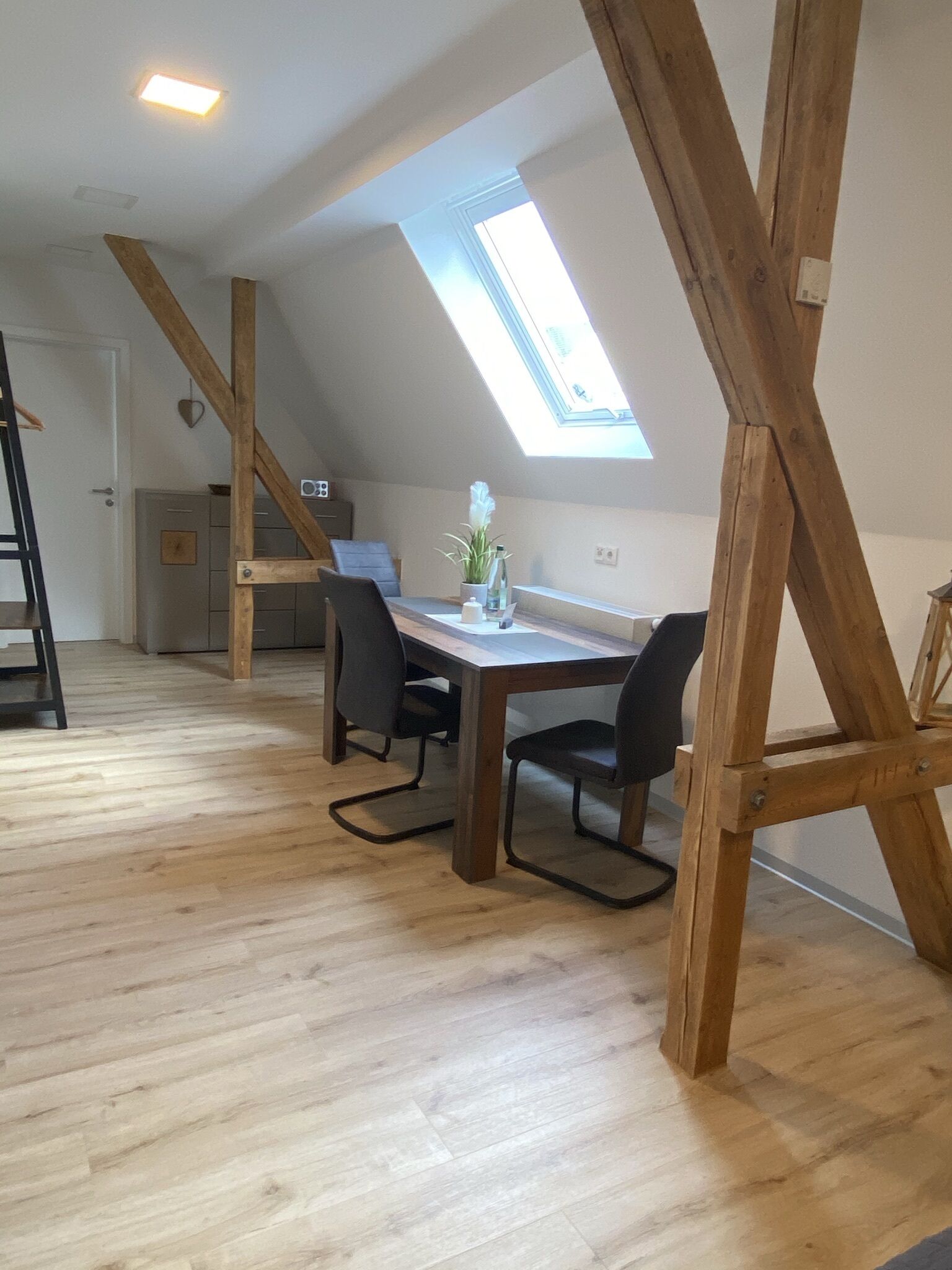 Ein Loft-Zimmer mit schräg verlaufender Decke, Holzbalken, einem Oberlicht, einem Esstisch mit Stühlen und einer kleinen Küchenzeile im Hintergrund.