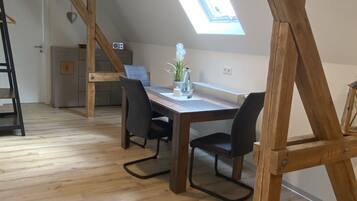 Ein Loft-Zimmer mit schräg verlaufender Decke, Holzbalken, einem Oberlicht, einem Esstisch mit Stühlen und einer kleinen Küchenzeile im Hintergrund.