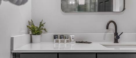 A modern bathroom with a white sink, a black faucet, and a mirror above it. There are three spa products on the counter and a potted plant to the left.