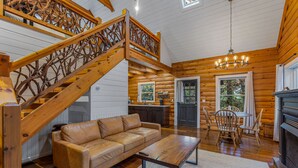 A log cabin interior with a wooden staircase, a leather sofa, a dining table with chairs, and a fireplace. - The Wells Hotel Cashiers (Cashiers)