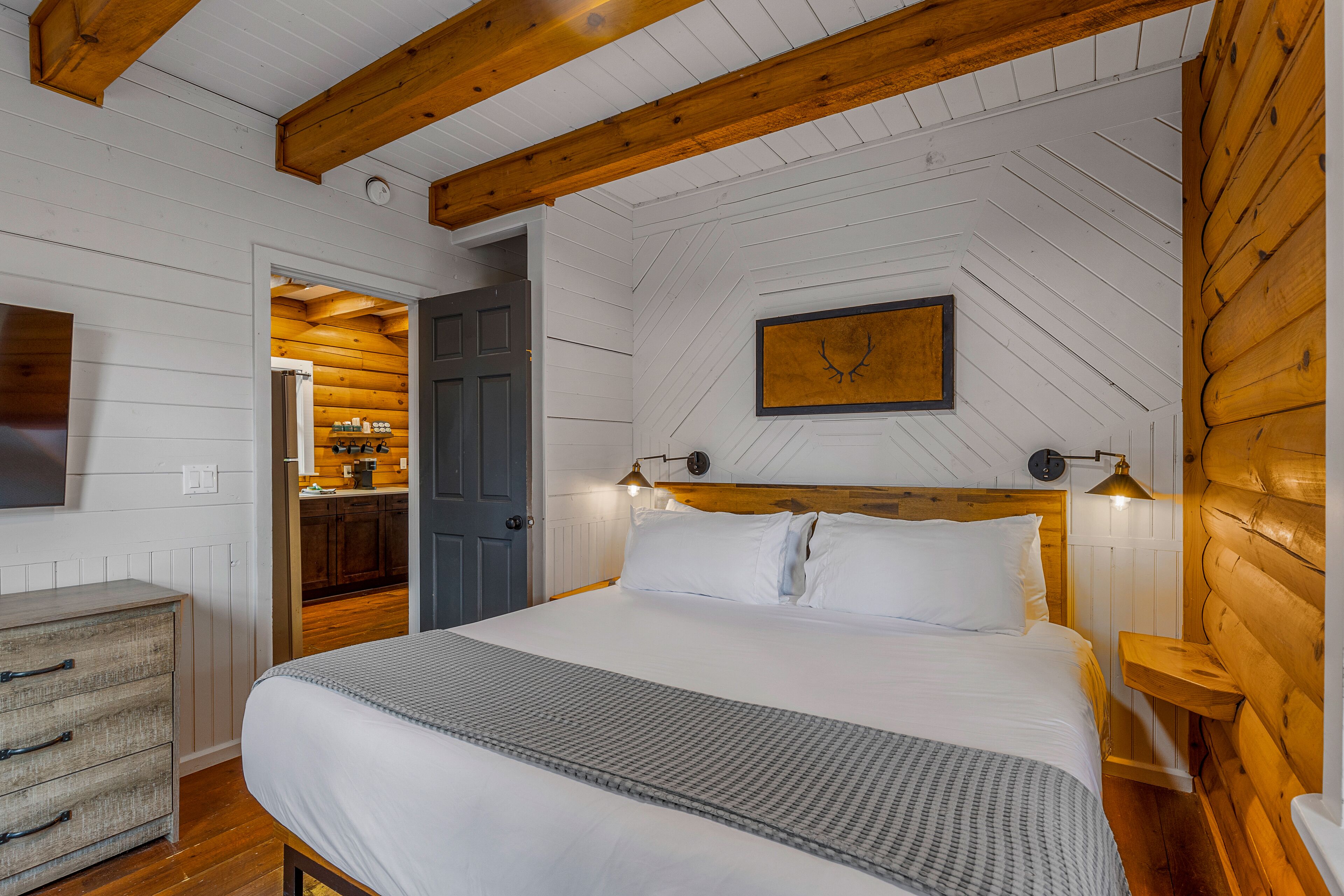 A bedroom with a wooden ceiling, a bed with white bedding, a bedside table, and a wall-mounted picture.