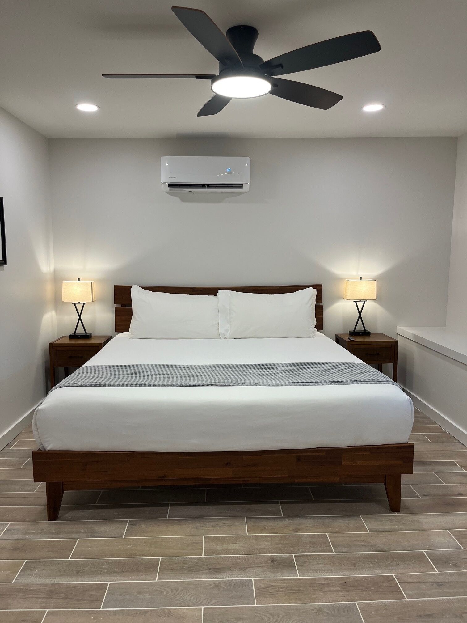 A neatly made bed with a wooden headboard, two bedside tables with lamps, a ceiling fan, and an air conditioning unit.