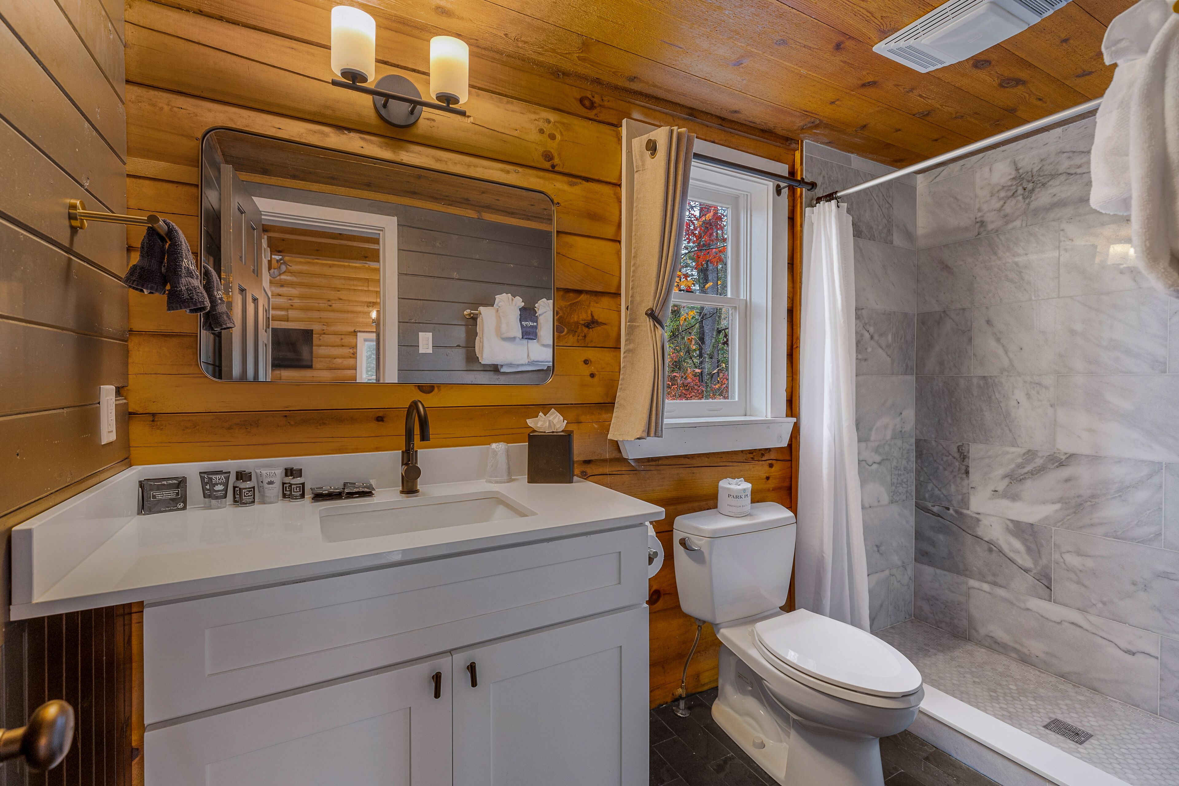 A bathroom with a wooden interior, a white sink, a mirror, a toilet, and a shower with a marble wall.