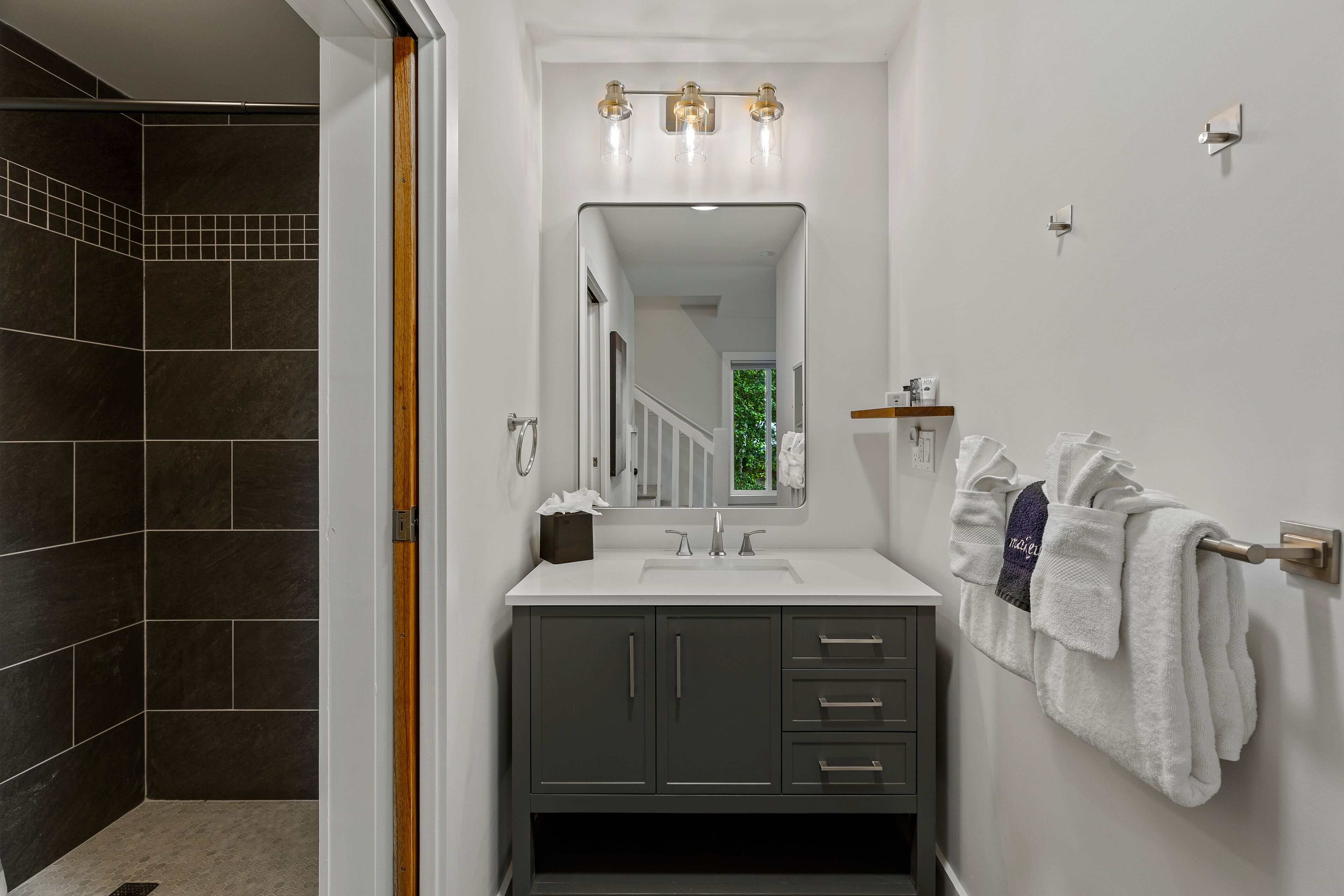 Een badkamer met een wit wastafelmeubel, donkergrijze kastjes, een spiegel met verlichting en een handdoekrek met handdoeken.