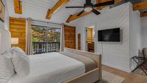 A bedroom with a large bed, wooden ceiling beams, a ceiling fan, a TV mounted on the wall, and a balcony with a view of trees. - The Wells Hotel Cashiers (Cashiers)